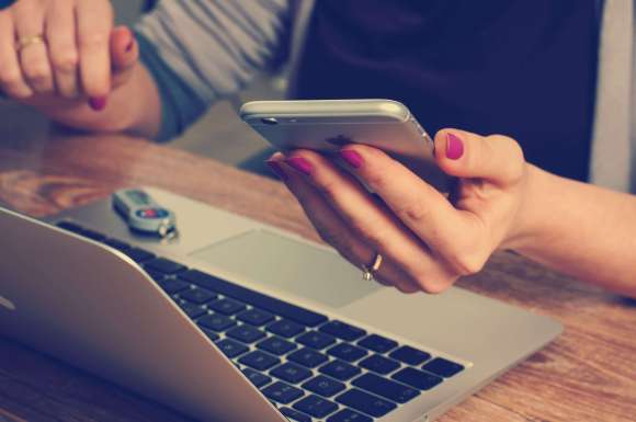 woman sat at laptop computer with mobile phone in hand