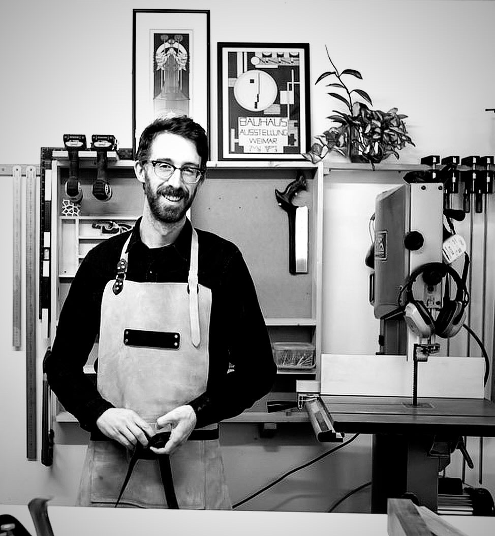 Cabinet maker smiles at camera standing in front of a shelf of tools in a workshop